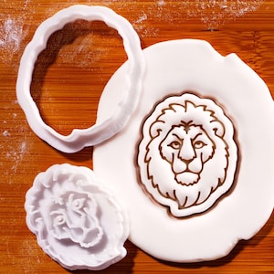 May include: A white lion-shaped cookie cutter and a lion-shaped fondant imprint on a wooden surface. The fondant imprint is brown and white. The cookie cutter is a wavy circle shape.