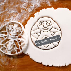 以下が含まれることがあります： 白いプラスチック製のクッキー型と、笑顔のキャラクターデザインのクッキー。クッキー型は、小麦粉をまぶした木製の表面にクッキーの隣にあります。クッキーには「FULL DETAILS IN LISTING」という文字が入っています。