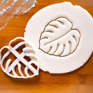 Könnte beinhalten: Ein weißer Ausstecher in Form eines Monstera-Blattes liegt neben einem weißen Teigausschnitt desselben Blattes. Der Ausstecher befindet sich auf einer Holzoberfläche mit einer Bestäubung aus weißem Mehl.