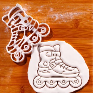 May include: A white plastic roller skate cookie cutter next to a roller skate-shaped cookie on a wooden surface. The cutter is a detailed outline of a roller skate, and the cookie is an imprint of the same design.