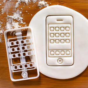 Pode incluir: Cortador de biscoitos de plástico branco em forma de smartphone com uma grade de botões na tela. O cortador está em uma superfície de madeira com açúcar em pó branco.