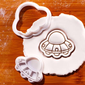 Könnte beinhalten: Auf einer Holzoberfläche werden weiße Kunststoff-Ausstecher in Form einer Wolke und eines UFOs zusammen mit einem Stück weißen Teigs gezeigt. Der UFO-Ausstecher wurde verwendet, um das Design in den Teig zu prägen. Ein Wolkenausstecher befindet sich in der Nähe.
