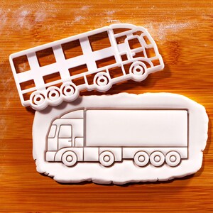 May include: A white plastic cookie cutter in the shape of a double-decker bus sits above a cookie cut in the shape of a semi-truck. The cookie cutter is on a wooden surface.