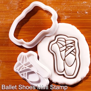 Peut inclure: Un emporte-pièce en plastique blanc et un tampon assorti représentant des chaussons de danse. Le tampon est pressé dans un morceau de pâte blanche circulaire, créant le contour de deux chaussons avec rubans. Texte : Ballet Shoes Mini Stamp.