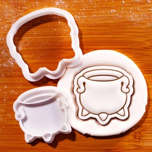 May include: White plastic cookie cutters in the shape of a cauldron and a witch's hat, along with a cauldron-shaped cookie. The cauldron cookie has a brown outline. The items are on a wooden surface.