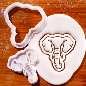 May include: White plastic elephant-shaped cookie cutters and a fondant elephant design. The cutters are in the shape of an elephant's head and body. The fondant elephant is brown and white.