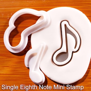 Op de afbeelding: Een witte plastic koekjesvorm in de vorm van een muzieknoot met bijbehorende stempel, waarbij een nootvorm in een rond stuk wit deeg is gedrukt. De tekst "Single Eighth Note Mini Stamp" staat in het wit onderaan de afbeelding gedrukt.