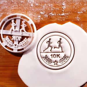 May include: White plastic cookie cutter featuring a running figure design, the text "10K", and laurel leaves. A matching imprint is visible on a white dough surface. The cutter is round with a cross-shaped design.
