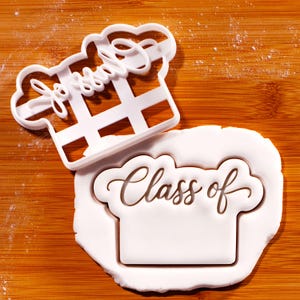 May include: White plastic cookie cutter in the shape of a chef's hat with the word "Jesse" and a grid design. A second cookie cutter in the shape of a chef's hat with the words "Class of" is on a white dough surface. The background is a wooden cutting board.