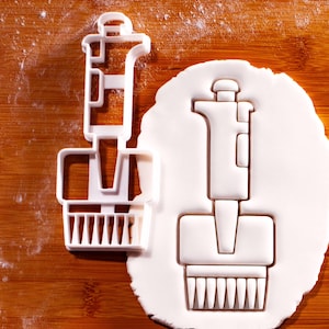 Könnte beinhalten: Ein weißer Ausstecher in Form einer Laborpipette, neben einem Keks in der gleichen Form. Der Ausstecher liegt auf einer Holzoberfläche mit Mehl. Die Pipettenform umfasst einen Griff, einen Körper und eine Spitze mit einem Raster.