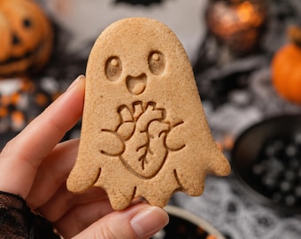 May include: A light brown, ghost-shaped biscuit featuring an embossed design of a smiling face and a detailed anatomical heart held in the centre. The biscuit has a smooth, baked texture with visible speckles throughout its surface.