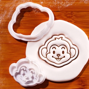 Könnte beinhalten: Weißer Kunststoff-Keks-Ausstecher in Affenform und ein Keks in Affenform auf einer Holzoberfläche. Der Ausstecher hat die Form einer Wolke. Der Affen-Keks hat ein lächelndes Gesicht.