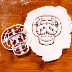May include: A white plastic cookie cutter shaped like a snake head, positioned next to a piece of white rolled dough that has been imprinted with the same snake head design, showing the detailed facial features of the reptile.