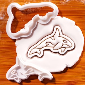 Op de afbeelding: Witte, orka-vormige koekjessnijders en een geperst orka-ontwerp in witte deeg. Het orka-ontwerp is bruin omlijnd. De uitstekers liggen op een houten oppervlak.