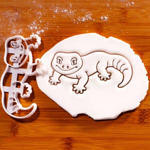 May include: A white plastic cookie cutter shaped like a gecko, positioned next to a piece of rolled-out white dough that has been imprinted with the same gecko design, featuring a smiling face, four legs, and a curved tail in the outline.