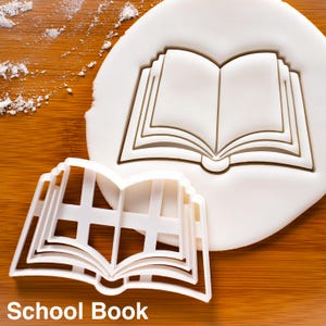 May include: A white plastic cookie cutter shaped like an open book, alongside an imprint of the same book shape in white dough. The text "School Book" is at the bottom of the image. The background is a wooden surface.