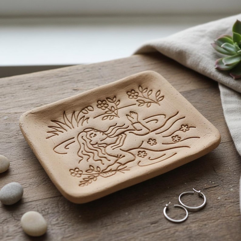 May include: A rectangular, light brown ceramic soap dish with an engraved design of a woman swimming in water, surrounded by flowers and plants. Two small silver hoop earrings are next to the dish.