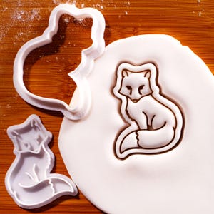 Könnte beinhalten: Ein weißer Ausstechform aus Kunststoff in Fuchsform und ein passender Stempel für Fondant. Eine Form ist leer abgebildet, während die andere in ein rundes Stück weißen Teigs gedrückt wurde, wodurch eine detaillierte Fuchskontur mit Gesichtszügen entsteht.