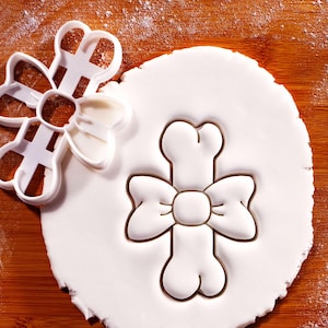 May include: A white plastic bow-shaped cookie cutter and a bow-shaped imprint in white dough. The cutter sits on a wooden surface dusted with flour. The dough imprint shows a detailed bow design.