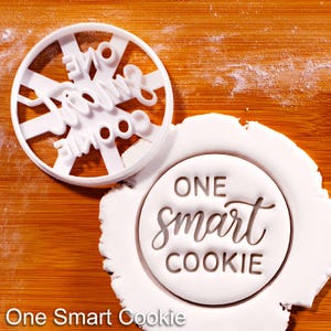 May include: A white plastic cookie cutter with the words "One Smart Cookie" in a circular design. The cutter is next to a round cookie with the same text imprinted. The words "One Smart Cookie" are also at the bottom of the image.