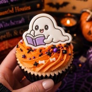 Peut inclure: Un cupcake surmonté d'un glaçage orange, de vermicelles noirs et violets, et de petits bonbons blancs en forme d'os. Un biscuit blanc en forme de fantôme souriant est posé dessus, tenant un livre violet ouvert avec des contours noirs.