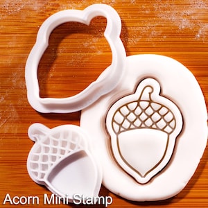 Könnte beinhalten: Weiße, eichelförmige Ausstecher und Stempel aus Kunststoff auf einer Holzoberfläche. Der Stempel hat ein detailliertes Eicheldesign. Der Text "Acorn Mini Stamp" ist unten im Bild zu sehen. Ideal zum Backen und Basteln.