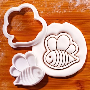 May include: White plastic bee-shaped cookie cutters and a bee-shaped fondant design on a wooden surface. The cutters include a bee outline, a cloud shape, and a bee with wings. The fondant design features a smiling bee with striped body.