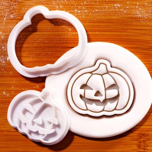 May include: White plastic pumpkin-shaped cookie cutters and a pumpkin-shaped cookie on a wooden surface. The cookie cutter has a round outer edge and a detailed pumpkin face design. The cookie is white with a brown outline.