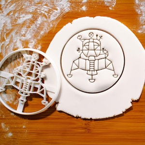 Può includere: Un tagliabiscotti in plastica bianca e un biscotto con un disegno in rilievo del modulo lunare Apollo. Il tagliabiscotti è a sinistra e il biscotto a destra, entrambi su una superficie di legno con farina.