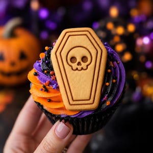 May include: A Halloween-themed cupcake featuring a light brown, coffin-shaped biscuit topper with an embossed skull design. The cupcake is decorated with swirls of vibrant orange and deep purple frosting, accented with small black and orange sprinkles.