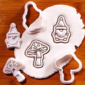 May include: A set of white plastic cookie cutters featuring two gnome shapes and two mushroom shapes. Some cutters are pressed into a circular piece of white dough, while others are placed separately, showing the outlines of the designs.