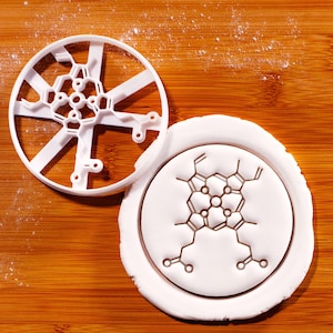 Op de afbeelding: Een witte plastic koekjesvorm in de vorm van een moleculaire structuur, naast een witte cirkel van deeg met hetzelfde ontwerp. De achtergrond is een houten oppervlak.