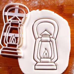 May include: A white plastic lantern-shaped cookie cutter next to a lantern-shaped imprint in white dough. The cutter has a handle and a flame design. The dough imprint shows the lantern's details.