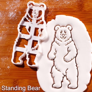 Pode incluir: Um cortador de biscoitos de plástico branco em forma de urso ao lado de um biscoito em forma de urso em uma superfície de madeira. O cortador é um recorte de um urso em pé. O biscoito é branco e tem um design detalhado de urso.
