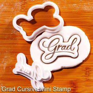 May include: White plastic cookie cutters and stamp on a wooden surface. The stamp says "Grad" in a cursive font. The text "Grad Cursive Mini Stamp" is at the bottom of the image.