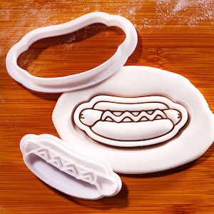 Può includere: Set di formine e timbro per biscotti a forma di hot dog in plastica bianca su una superficie di legno. Il set include una formina per il panino, un timbro con il disegno dell'hot dog e della senape e una formina più piccola per la forma dell'hot dog.