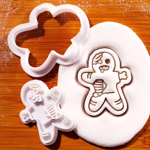 以下が含まれることがあります： 白いプラスチック製のクッキー型で、雲とゾンビのジンジャーブレッドマンの形をしています。ゾンビのジンジャーブレッドマンが白い生地に押し込まれています。この型は、ハロウィーンや不気味なテーマのクッキーを作るためのものです。