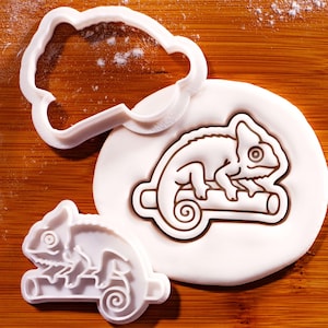 May include: A white plastic chameleon cookie cutter and a matching stamp, shown alongside a piece of white dough imprinted with the chameleon design. The design features a chameleon perched on a branch with a curled tail and a detailed eye.