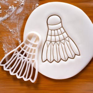 Könnte beinhalten: Ein weißer Ausstecher in Form eines Federballs neben einem Federball-Abdruck auf weißem Fondant. Der Ausstecher befindet sich auf einer Holzoberfläche mit einer Staubschicht aus weißem Pulver.