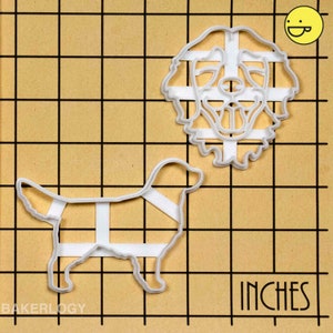 May include: Two white plastic cookie cutters in the shape of a golden retriever dog. The larger cutter is a head and the smaller cutter is the full body of the dog. The cutters are on a brown gridded surface with the word "INCHES" printed on the bottom right corner.