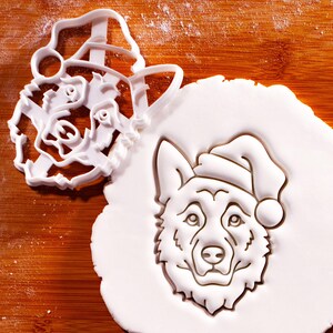 May include: A white plastic cookie cutter in the shape of a dog's head wearing a Santa hat, next to a cutout of the same design in white dough. The dog's face is detailed with eyes, nose, and fur.