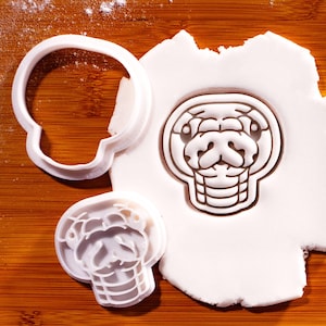 May include: Two white plastic cookie cutters shaped like a snake head. One cutter is pressed into a piece of white dough, creating a detailed impression of the snake's face, including eyes, nostrils, and scales. The other cutter sits nearby.