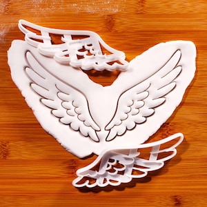May include: White angel wing cookie cutters and a wing-shaped fondant design on a wooden surface. The cutters are in different sizes, with detailed feather impressions. The fondant has a detailed wing design, perfect for baking and crafting.