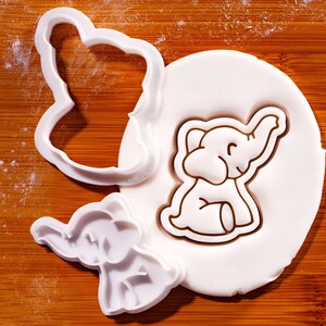 May include: White plastic elephant-shaped cookie cutters and a fondant elephant on a wooden surface. The fondant elephant is outlined in brown. One cutter is the outline of an elephant, and the other is the shape of the elephant.