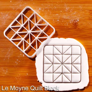 Può includere: Un tagliabiscotti Le Moyne Quilt Block in plastica bianca accanto a un disegno di blocco trapuntato pressato in pasta bianca. Il tagliabiscotti è un quadrato con un motivo geometrico. Il testo "Le Moyne Quilt Block" è in basso.