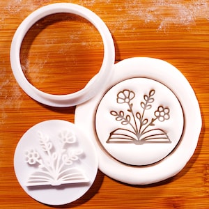 Op de afbeelding: Witte koekjessnijderset met een bloemen- en boekontwerp. De set bevat een ronde uitsteker en twee stempels met een bloem- en boekillustratie. Het ontwerp is in een witte cirkel geperst.