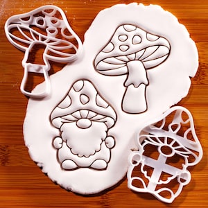 May include: Two white plastic cookie cutters shaped like a mushroom and a gnome with a mushroom hat, alongside impressions of the same designs pressed into a flat piece of white dough, showing detailed outlines of the mushroom cap and gnome beard.