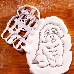 May include: A white cookie cutter and a white cookie shaped like a fluffy dog sitting down. The cookie cutter is in the shape of a bow. The background is a wooden cutting board.