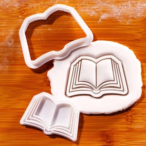 May include: White plastic cookie cutters shaped like an open book. One is an outline, another an impression in dough, and a third a smaller, formed book shape. The cutters are on a wooden surface.