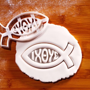 Puede incluir: Un cortador de galletas blanco con forma de pez con la palabra griega "ΙΧΘΥΣ" en su interior. El cortador se coloca sobre un trozo de masa blanco con forma de pez con la misma palabra impresa. El fondo es una superficie de madera.